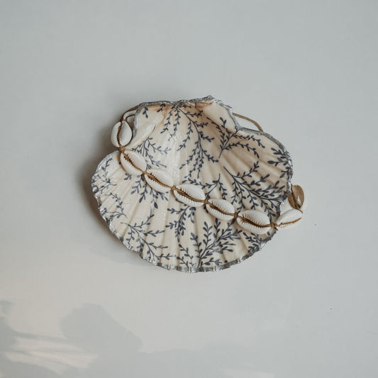 Handbemalte Jakobsmuschel mit blauem Blattmuster und silberfarbenem Rand, darin ein Armband aus weißen Kaurimuscheln auf braunem Band.