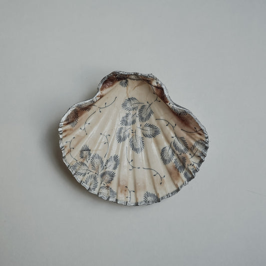 Handbemalte Jakobsmuschel mit silbernem Rand, cremefarbenem Grund und feinem schwarzen Blättermuster, auf hellem Hintergrund fotografiert.