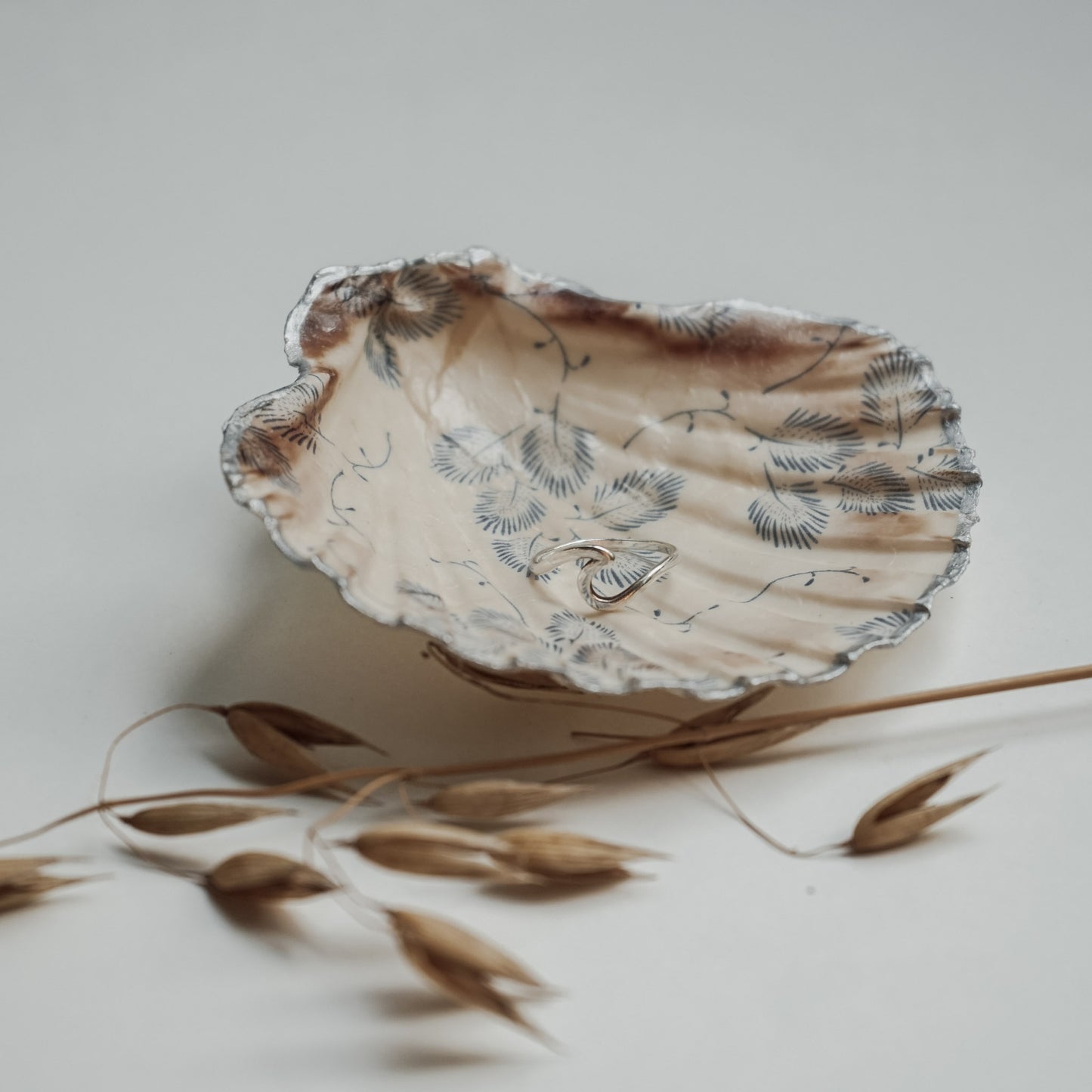 Handbemalte Jakobsmuschel mit silberfarbenem Rand und filigranem schwarz-grauem Blattmuster, genutzt als Schmuckschale mit einem silbernen Ring darin, dekoriert mit getrockneten Grashalmen auf hellem Hintergrund.