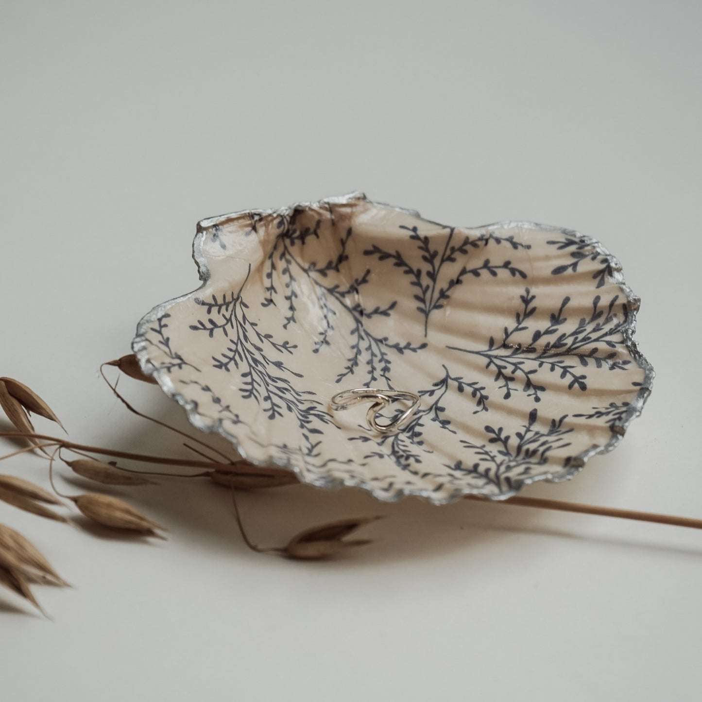 Handbemalte Jakobsmuschel mit blauem Blattmuster und silberfarbenem Rand, darin ein silberner Ring, daneben dekorative Trockenhalme auf hellem Untergrund.