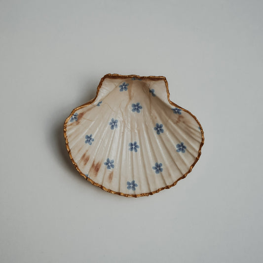 Handbemalte Jakobsmuschel mit cremefarbenem Hintergrund, gleichmäßig verteilten kleinen blauen Blüten und goldfarbenem Rand, flach auf hellem Untergrund liegend.