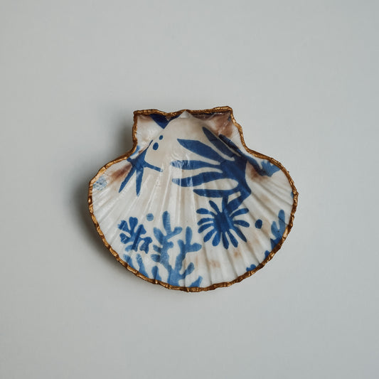 Handbemalte Jakobsmuschel mit goldfarbenem Rand und blauem Motiv aus maritimen Pflanzen und Korallen, auf hellem Hintergrund.