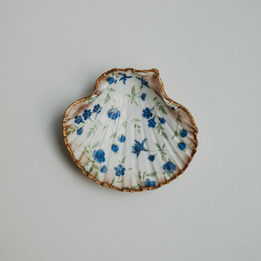 Handbemalte Jakobsmuschel mit goldfarbenem Rand und blau-grünem Blumenmuster, flach auf weißem Untergrund liegend.