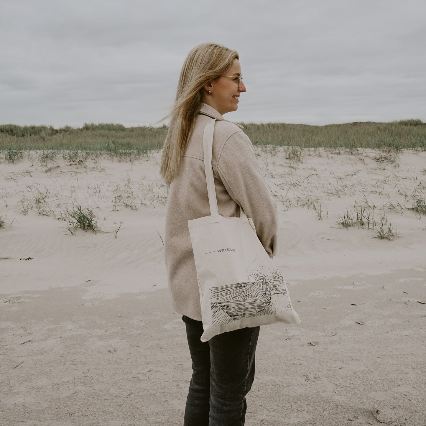 Frau mit langen blonden Haaren steht seitlich am Strand, trägt beigefarbene Jacke und eine helle Stofftasche mit Wellenmotiv und der Aufschrift 'WELLENLIEBE'; im Hintergrund Sanddünen und grauer Himmel.
