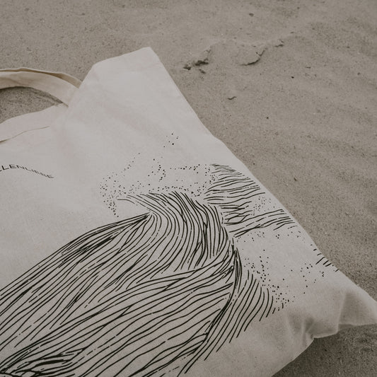 Beigefarbene Stofftasche liegt im Sand, bedruckt mit einer schwarzen, grafischen Wellenillustration und feinen Punkten, die Gischt darstellen.