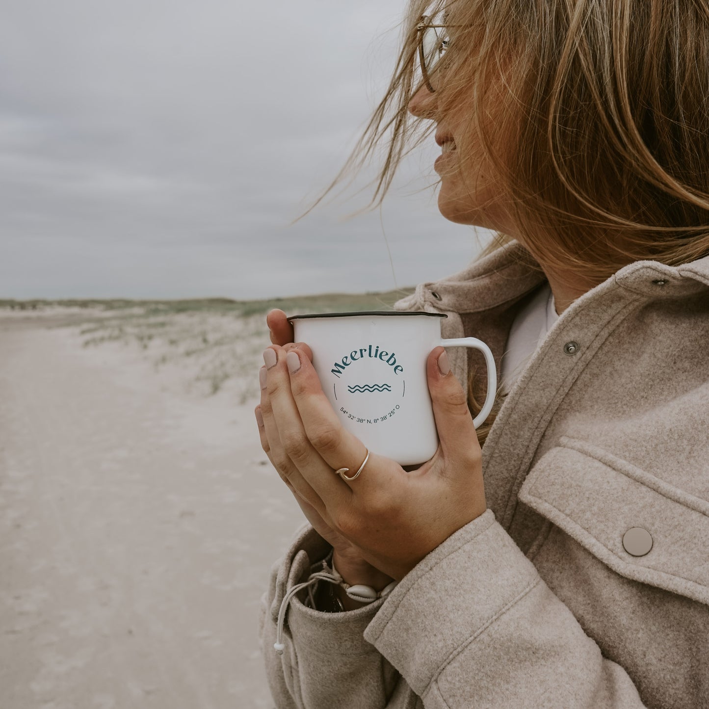 Frau mit offenen Haaren und beigem Mantel hält eine weiße Emaille-Tasse mit der Aufschrift 'Meerliebe' und Koordinaten in beiden Händen, lächelnd am Strand mit Dünen im Hintergrund.