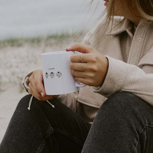Frau im beigefarbenen Mantel und dunkler Jeans sitzt am Strand und hält eine weiße Keramiktasse mit drei gezeichneten Schafen und dem Schriftzug 'määäh' in beiden Händen.
