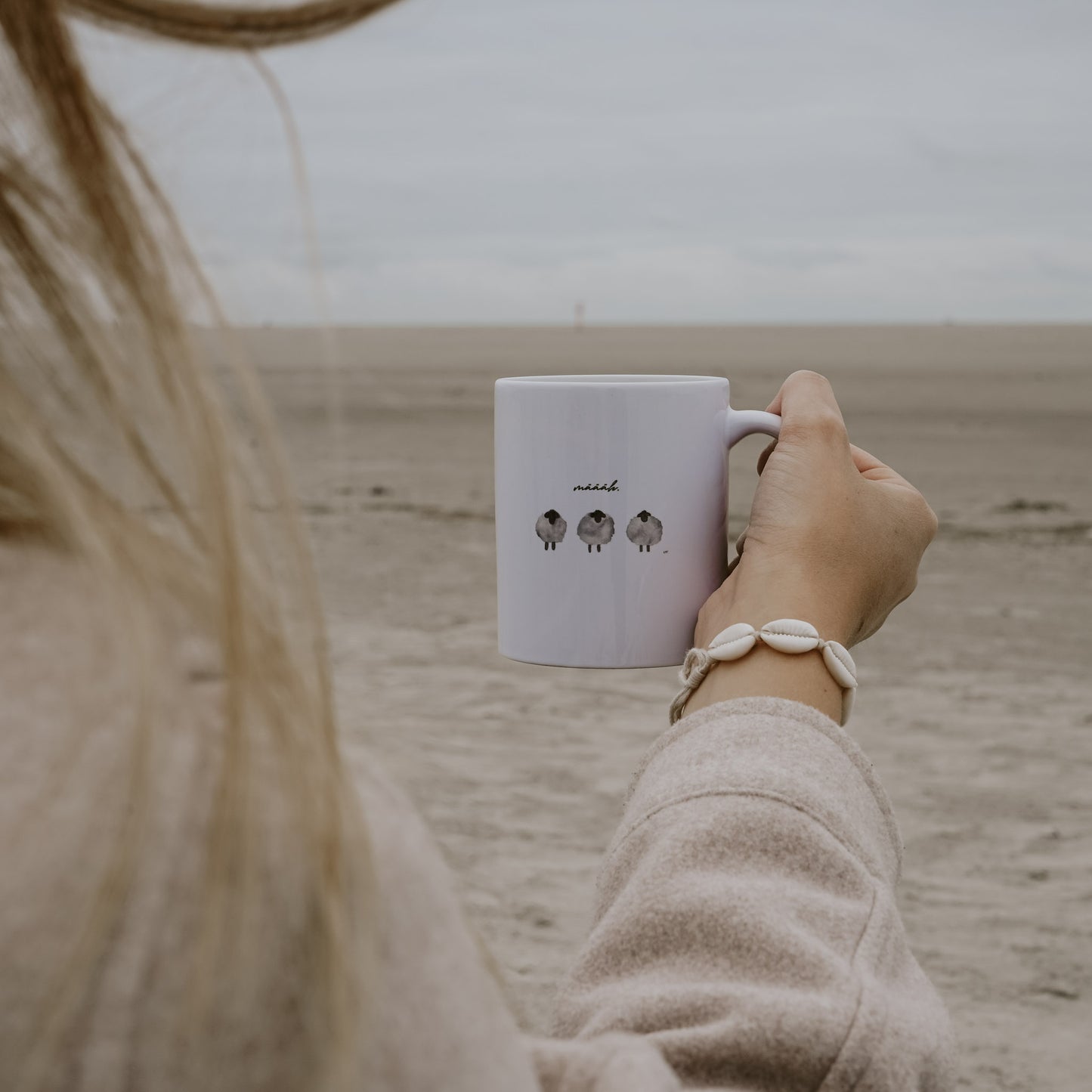 Person hält am Strand eine weiße Tasse mit drei gezeichneten Schafen und dem Schriftzug 'määäh.' in der Hand, trägt ein Armband aus Muscheln und eine beigefarbene Jacke.
