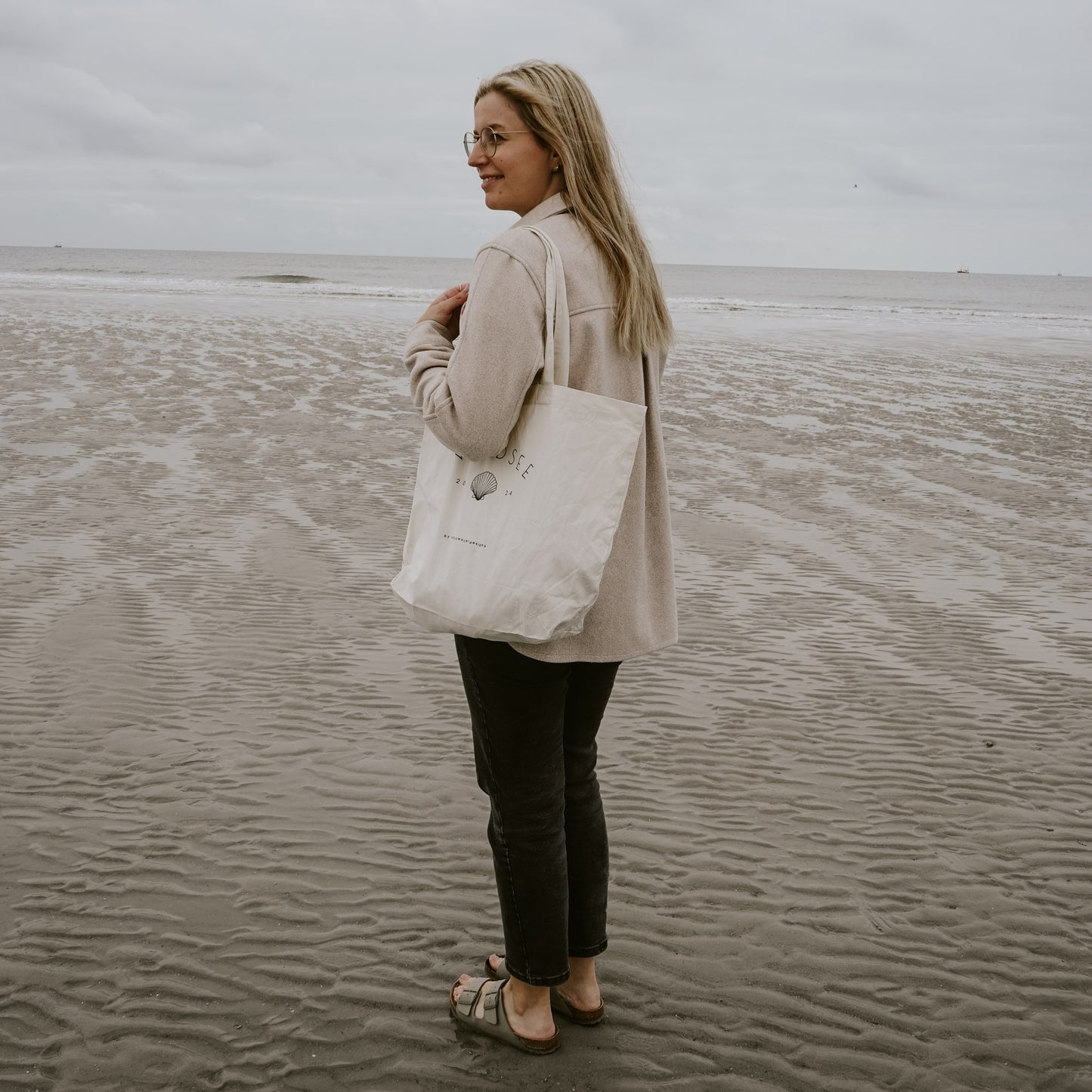 Frau mit blonden Haaren, Brille, beiger Jacke und schwarzer Hose steht barfuß in Sandalen am Strand, hält eine beigefarbene Stofftasche mit der Aufschrift 'Nordsee 2024' und einer Muschel-Illustration, im Hintergrund das Meer unter grauem Himmel.