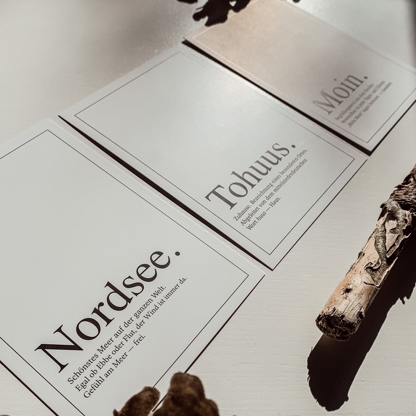 Drei minimalistische Postakarten mit den Worten „Nordsee.“, „Tohuus.“ und „Moin.“, seitlich versetzt auf einer hellen Fläche arrangiert, mit getrocknetem Holzdekor, fotografiert aus einem schrägen Winkel im warmen Sonnenlicht.