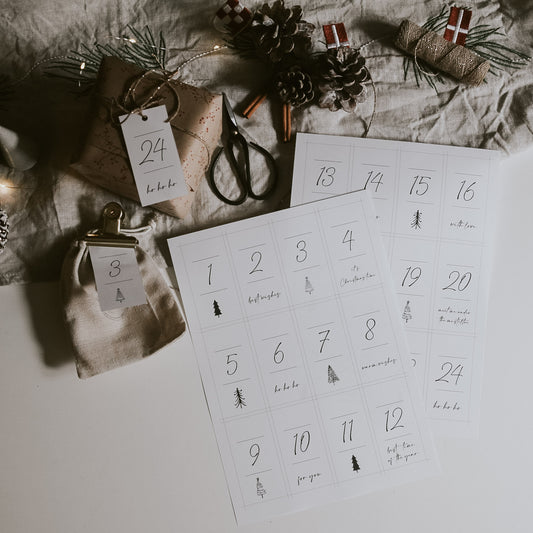 Zwei Bögen mit Adventskalender-Zahlenkarten im minimalistischen Design liegen auf einem Tisch, daneben ein verpacktes Geschenk mit Anhänger, eine kleine Leinen-Geschenktüte mit Klammer, eine Schere, Tannenzapfen und weihnachtliche Dekoration.