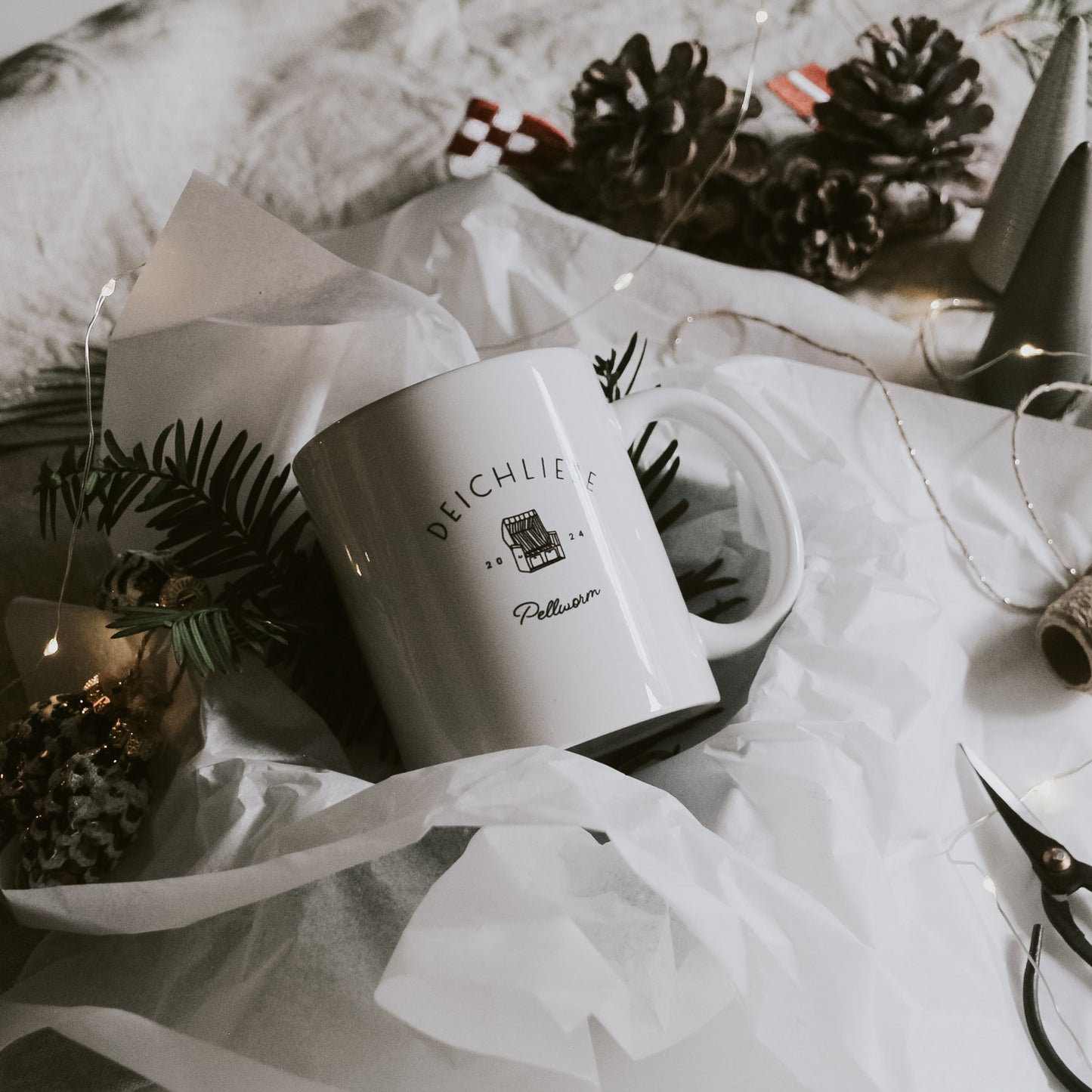 Weiße Keramiktasse mit der Aufschrift 'DEICHLIEBE Pellworm 2024' und einer Illustration eines kleinen Hauses, dekorativ in weißem Seidenpapier gebettet, umgeben von Tannenzweigen, Kiefernzapfen, goldenen Kugeln und einer Lichterkette, mit Schere und Bastelschnur im Hintergrund.