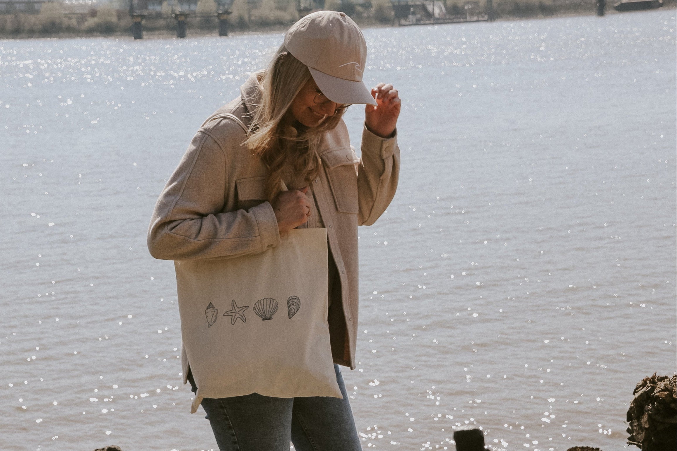 Junge Frau mit Cap und beigefarbener Jacke trägt eine Stofftasche mit Muschelmuster, steht entspannt am sonnigen Elbufer