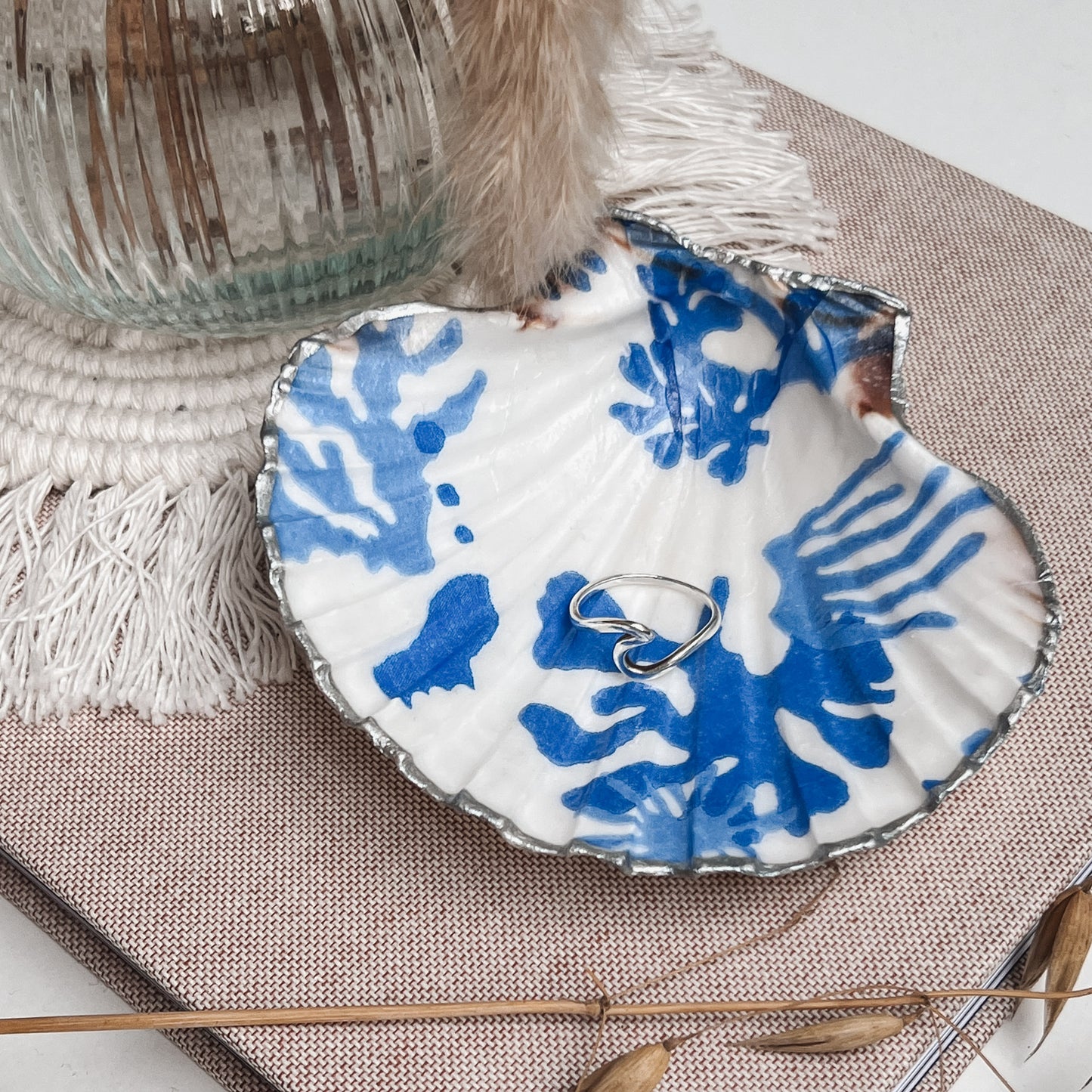 Handbemalte Jakobsmuschel mit silbernem Rand und blauem maritimem Muster, in der ein silberner Ring liegt, dekorativ arrangiert auf einer Stoffunterlage mit Glasvase, Makramee-Elementen und Trockenblumen.