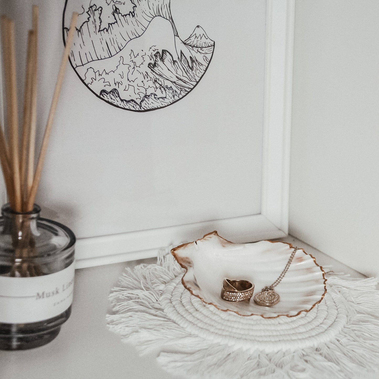 Weiße Jakobsmuschel mit goldfarbenem Rand, gefüllt mit einem goldenen Ring und einer goldenen Halskette mit rundem Anhänger, platziert auf einem geflochtenen weißen Untersetzer vor einem gerahmten Kunstwerk und neben einem Raumduft.