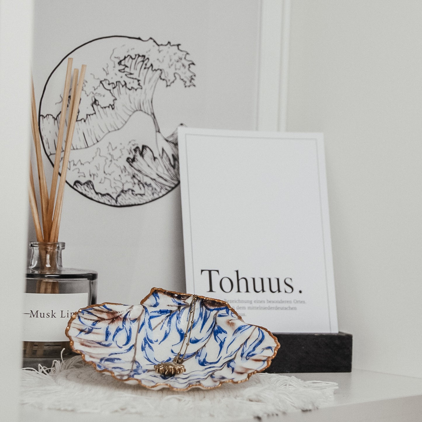 Handbemalte Muschelschale mit blauem Muster und goldfarbenem Rand, dekorativ platziert vor einem gerahmten Print mit dem Schriftzug 'Tohuus' und einem Glas-Duftdiffuser.