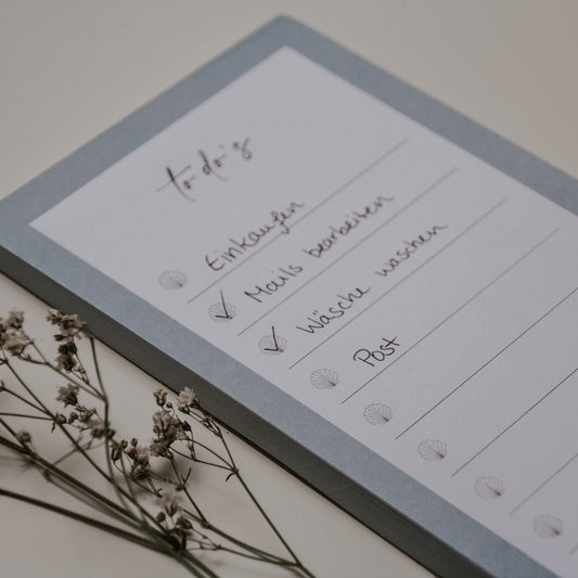 Nahaufnahme eines rechteckigen Notizblocks im Hochformat mit hellgrauem Rand und der Überschrift 'to-do's' in handschriftlicher Schriftart. Die Liste enthält die Einträge 'Einkaufen', 'Mails beantworten' (abgehakt), 'Wäsche waschen' (abgehakt) und 'Post'. Jede Zeile ist mit einer kleinen Muschel-Illustration als Aufzählungszeichen versehen. Links neben dem Block liegen kleine weiße Trockenblüten.