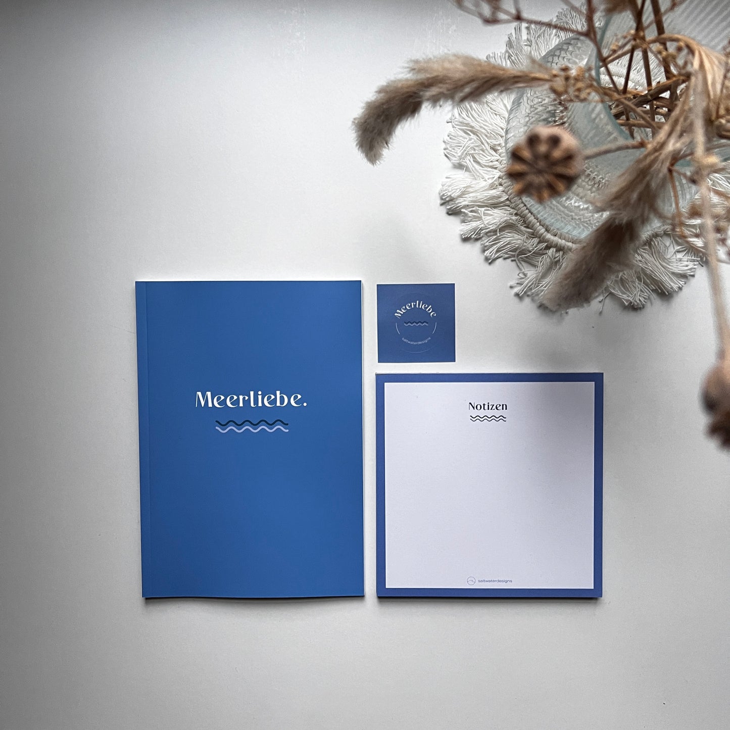 Flach gelegtes Set aus einem blauen Notizbuch mit der Aufschrift 'Meerliebe.', einem quadratischen Notizblock mit der Überschrift 'Notizen' und einem kleinen quadratischen Sticker, arrangiert auf einer hellen Oberfläche neben getrockneten Gräsern und einer weißen Makramee-Unterlage.