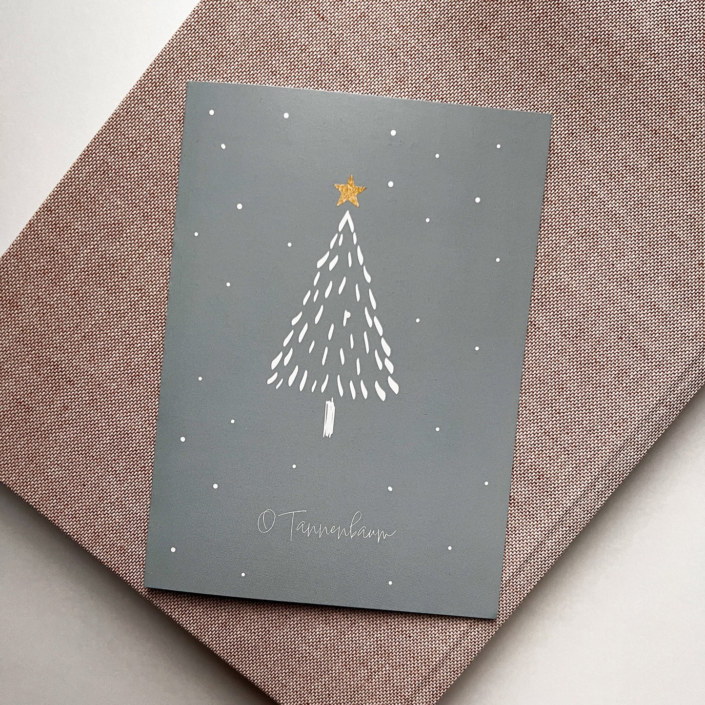 Weihnachtskarte mit minimalistischem Tannenbaum in Weiß auf grauem Hintergrund, verziert mit einem gelben Stern an der Spitze und kleinen weißen Punkten als Schneefall, platziert auf einem rosafarbenen Stoffuntergrund.