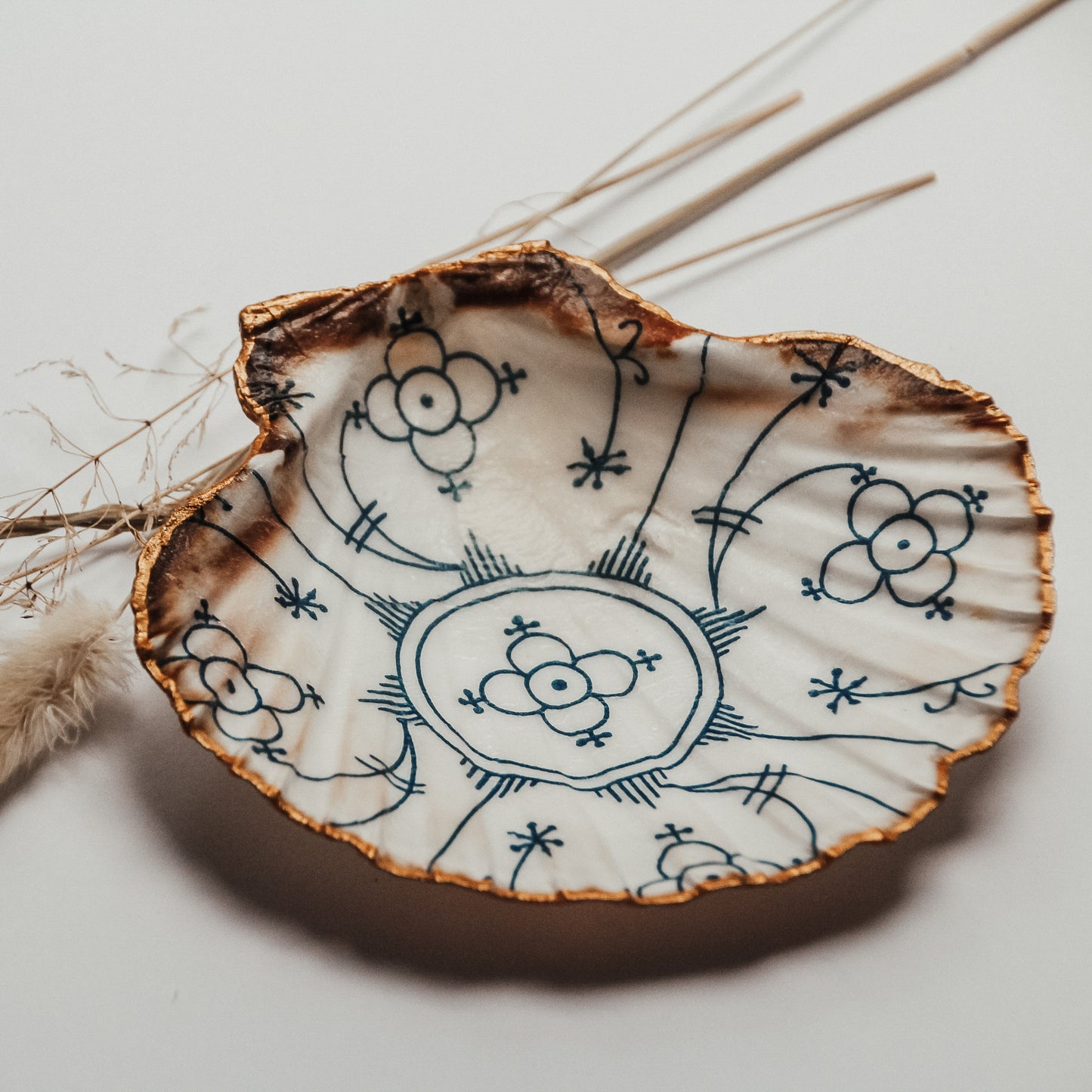 Handbemalte Jakobsmuschel-Schale mit blauem floralen Muster und goldenem Rand, liegend auf weißem Hintergrund mit getrockneten Gräsern im Hintergrund.