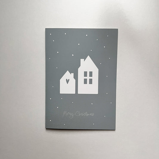 Vorderseite der grauen Weihnachtskarte mit zwei weißen Häusern und Schneeflocken in minimalistischem Design, mittig platziert vor neutralem Hintergrund.