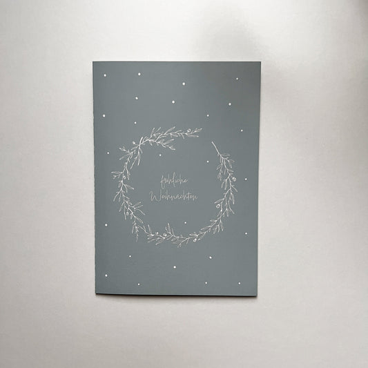 Minimalistische Weihnachtskarte in Grau mit weißem, filigranem Kranz-Design und der Aufschrift „frohe Weihnachten“, kombiniert mit weißem Umschlag auf hellem Hintergrund.
