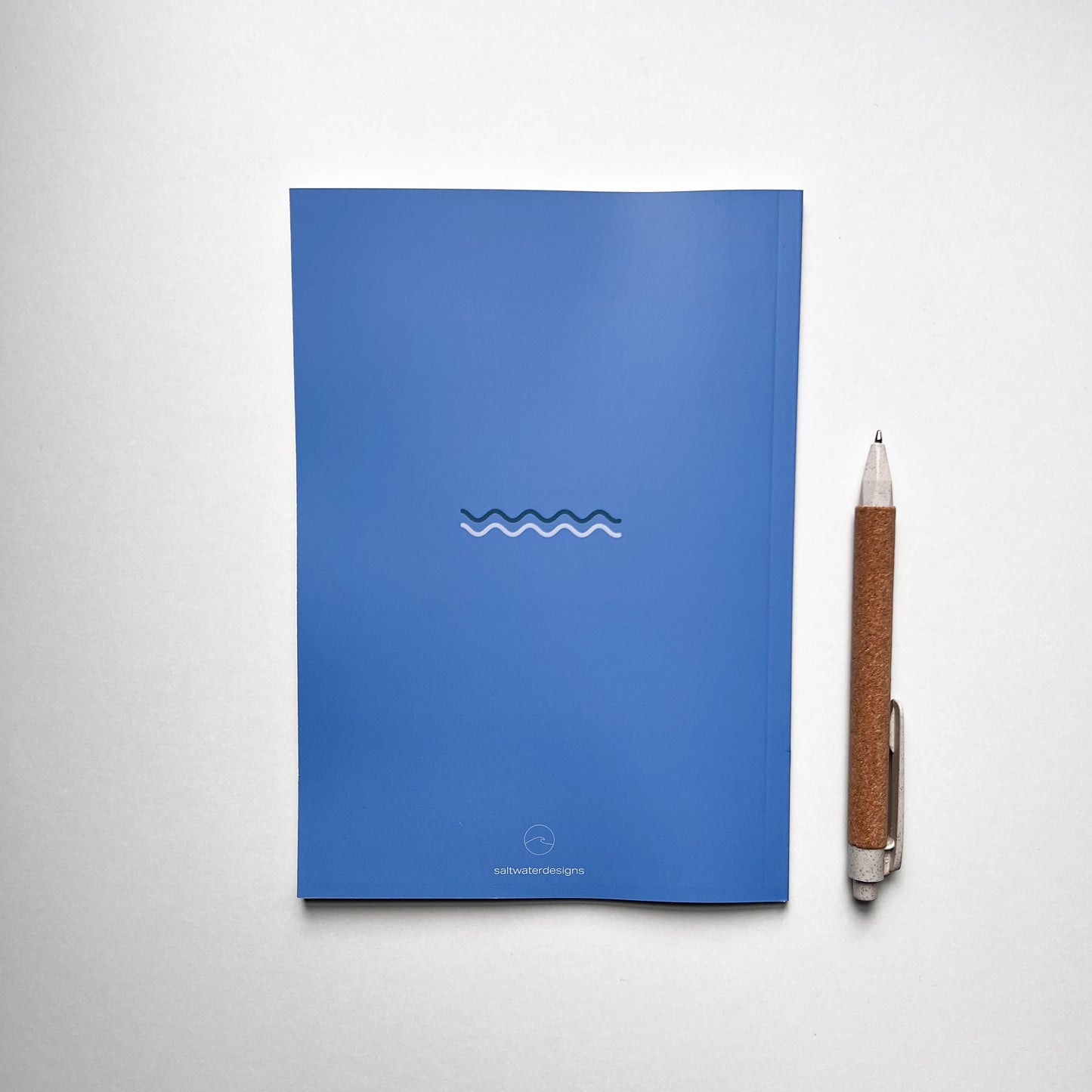 Blaues Notizbuch mit stilisierter Wellenlinie in der Mitte und kleinem 'saltwaterdesigns'-Logo unten. Daneben liegt ein Kugelschreiber mit brauner, korkähnlicher Oberfläche auf weißem Hintergrund.