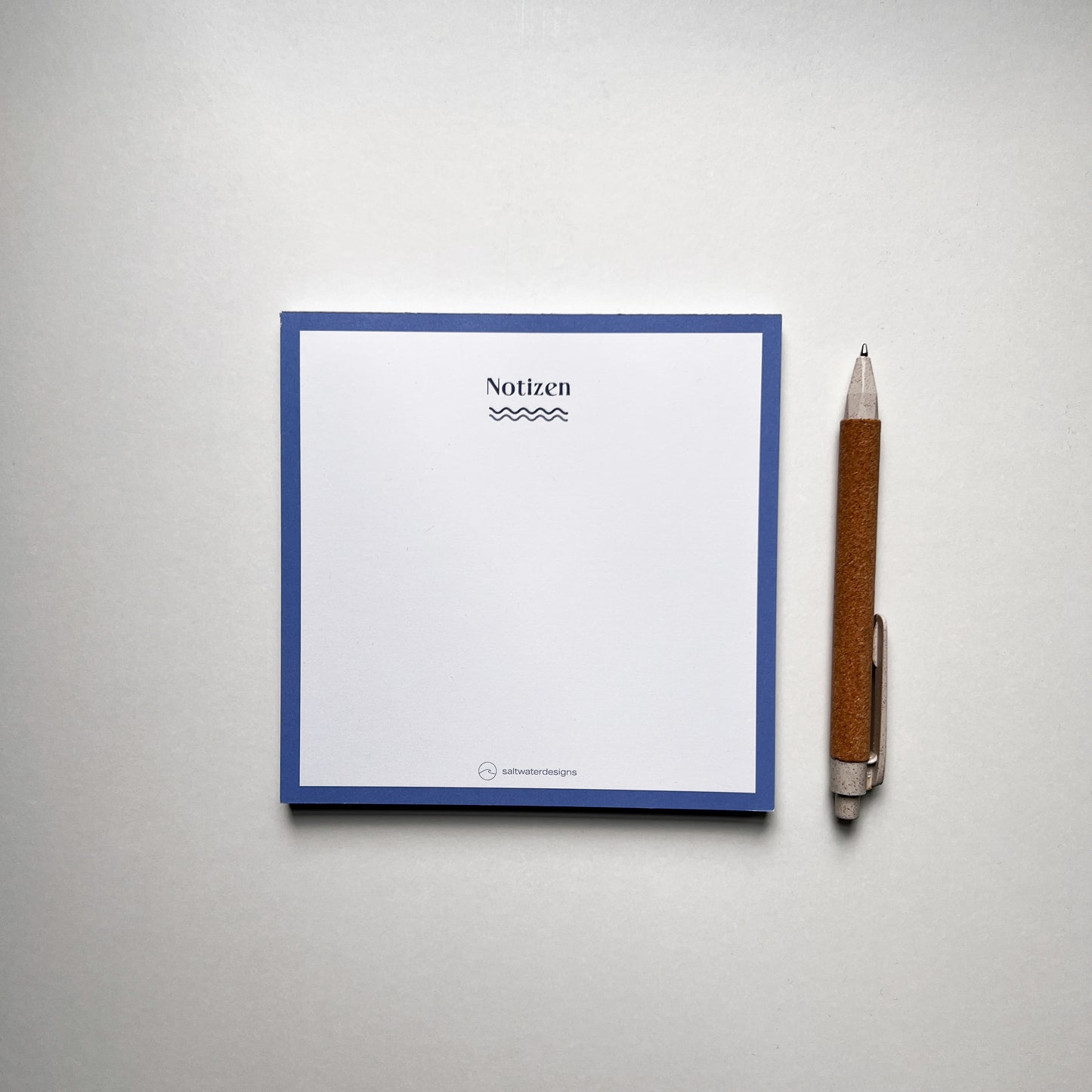 Quadratischer Notizblock mit blauem Rand und der Aufschrift 'Notizen' oben, darunter eine wellenförmige Linie. Unten mittig das Logo 'saltwaterdesigns'. Rechts neben dem Block liegt ein brauner Kugelschreiber mit silbernem Clip auf einer hellen Oberfläche.