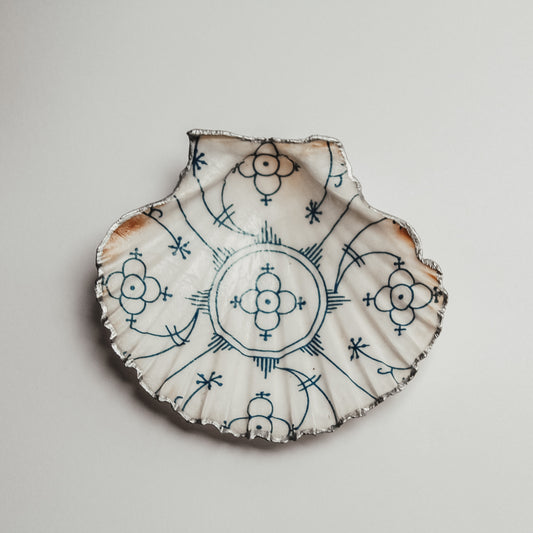 Handbemalte Muschelschale mit symmetrischem, blauem Ornamentmuster und silberfarbenem Rand, dekorativ auf hellem Untergrund platziert.