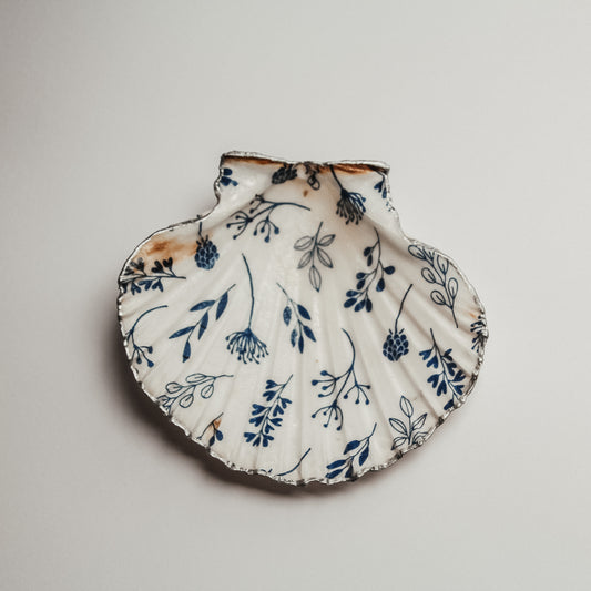 Weiße Jakobsmuschel mit silberfarbenem Rand und handgemaltem blauen Pflanzen- und Blütenmuster, auf hellem Untergrund fotografiert.