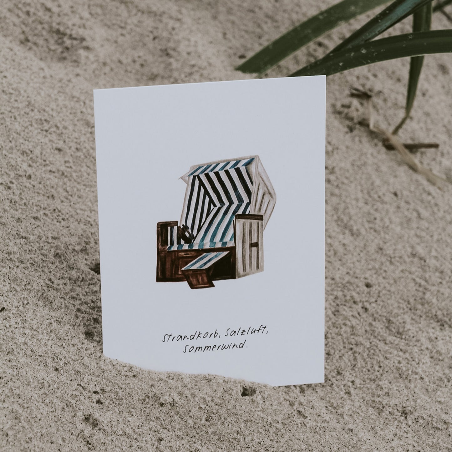 Postkarte mit maritimer Illustration: Ein einzelner Strandkorb steht im Sand vor einem hellen Himmel. Die Karte mit dem Schriftzug „Strandkorb, Salzluft, Sommerwind“ ist leicht in den Sand gesteckt und vermittelt ein sommerliches Nordsee-Flair.