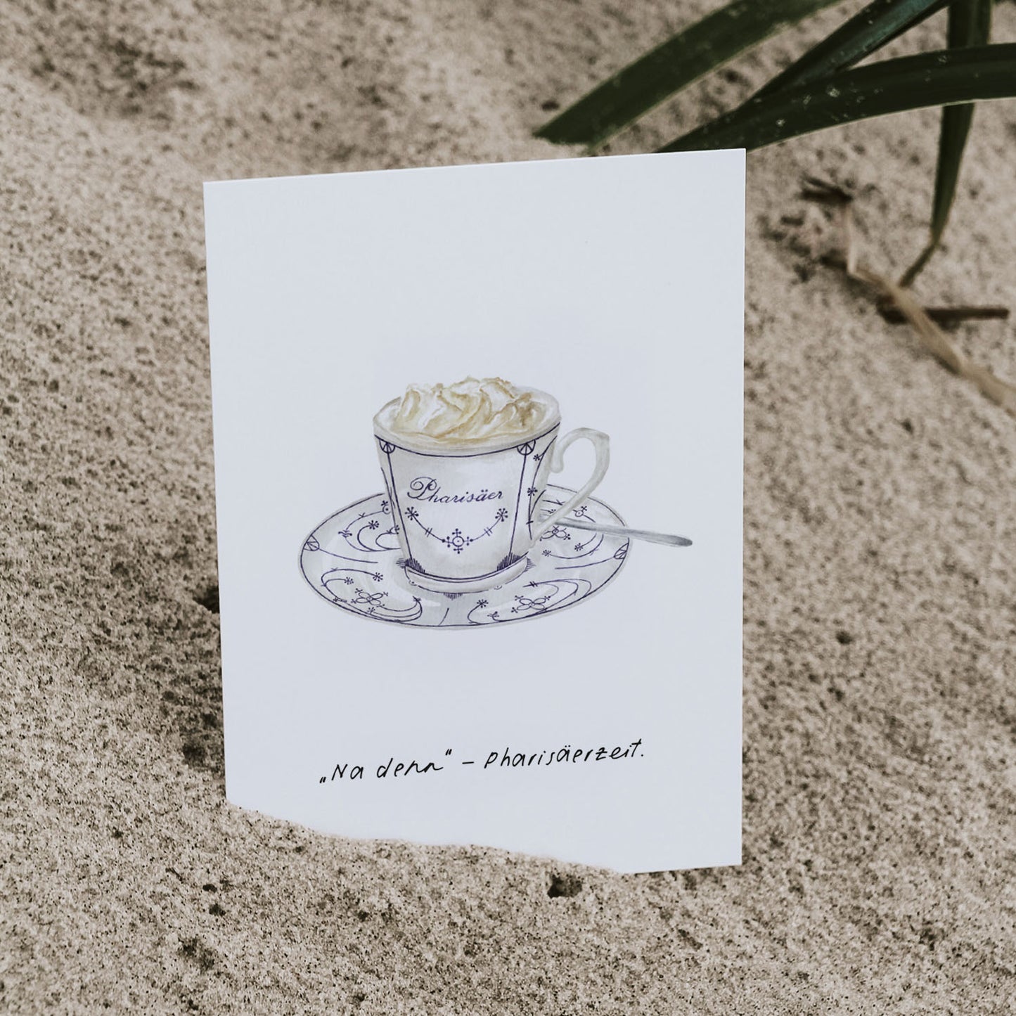 Nahaufnahme einer nordfriesischen Postkarte im Aquarellstil mit handgezeichneter Pharisäer-Illustration. Die Karte steckt leicht im Sand und zeigt den humorvollen Spruch 'Na denn – Pharisäerzeit'. Ein charmantes Geschenk für Nordsee-Fans.
