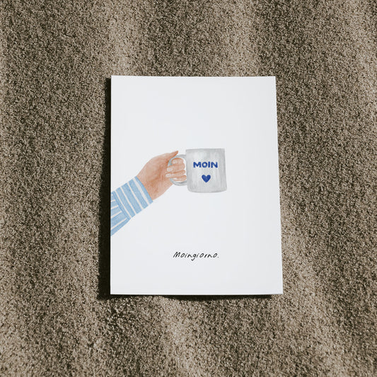 Postkarte mit Aquarellmotiv: Hand hält eine Kaffeetasse mit 'Moin'-Schriftzug. Darunter steht der Spruch 'Moingiorno'. Die Karte liegt dekorativ auf hellem Sand – norddeutscher Humor trifft italienische Strandatmosphäre."