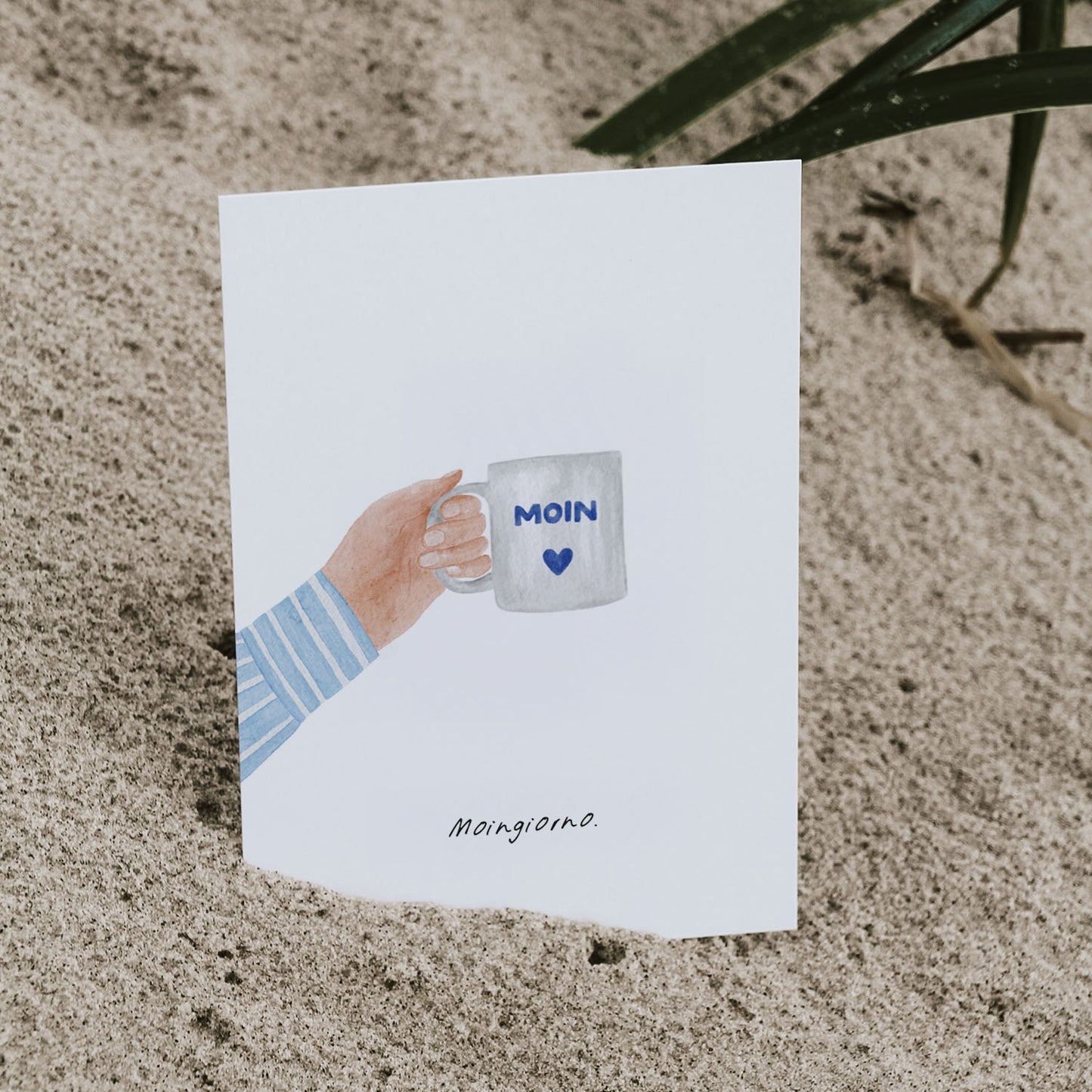 Postkarte im Aquarellstil, halb in Sand gesteckt: Hand mit 'Moin'-Tasse und dem Spruch 'Moingiorno'. Ideal für Fans von Kaffee, Küstenflair und originellen Wortspielen.