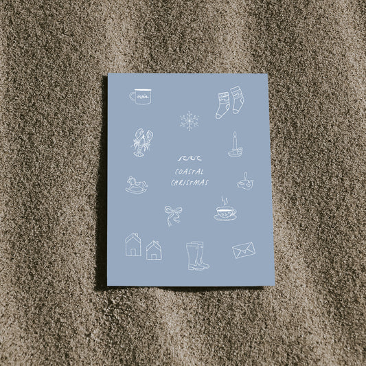 Blaue Postkarte mit dem Schriftzug „Coastal Christmas“ und nordischen, maritimen Illustrationen, die auf sandigem Untergrund liegt.