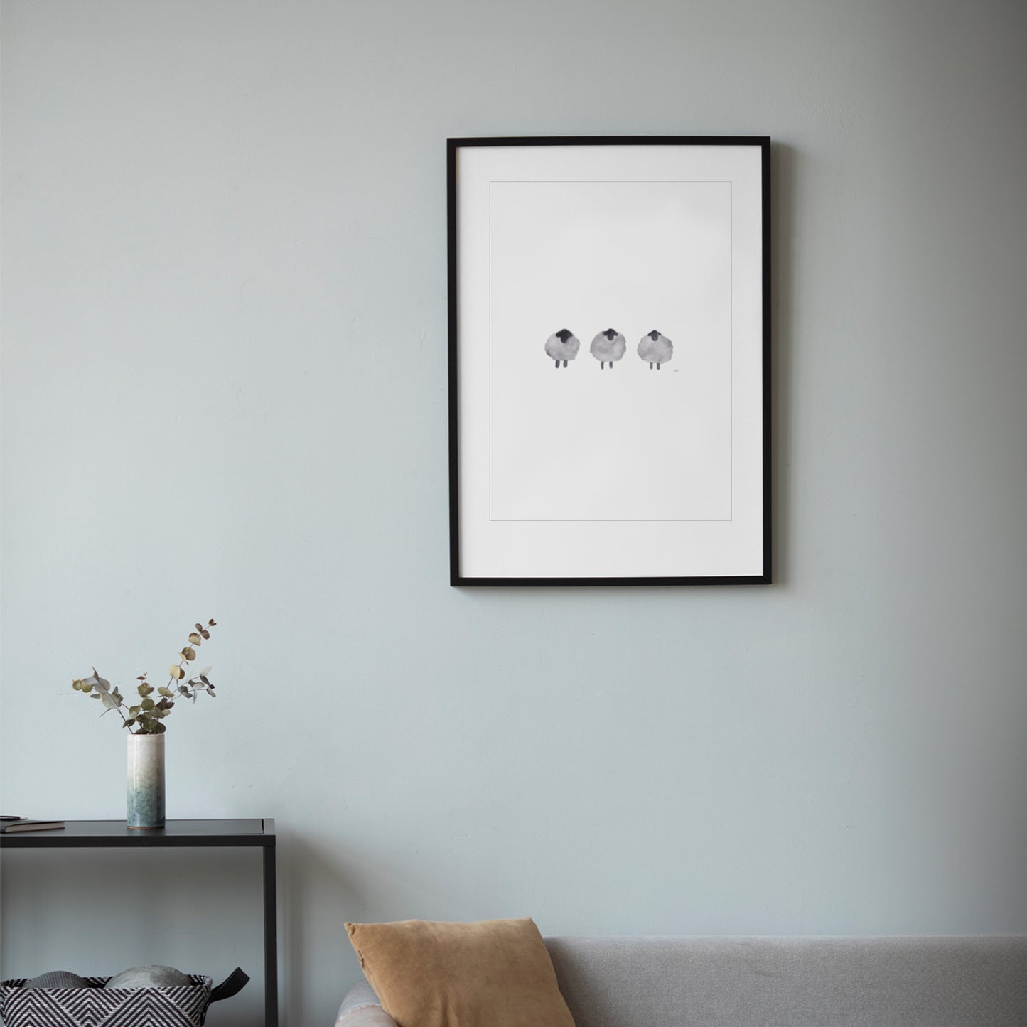 Minimalistisches Poster mit drei schwarzen Schafen in Aquarelloptik, im schwarzen Rahmen an einer hellgrauen Wand über einem Sofa hängend.