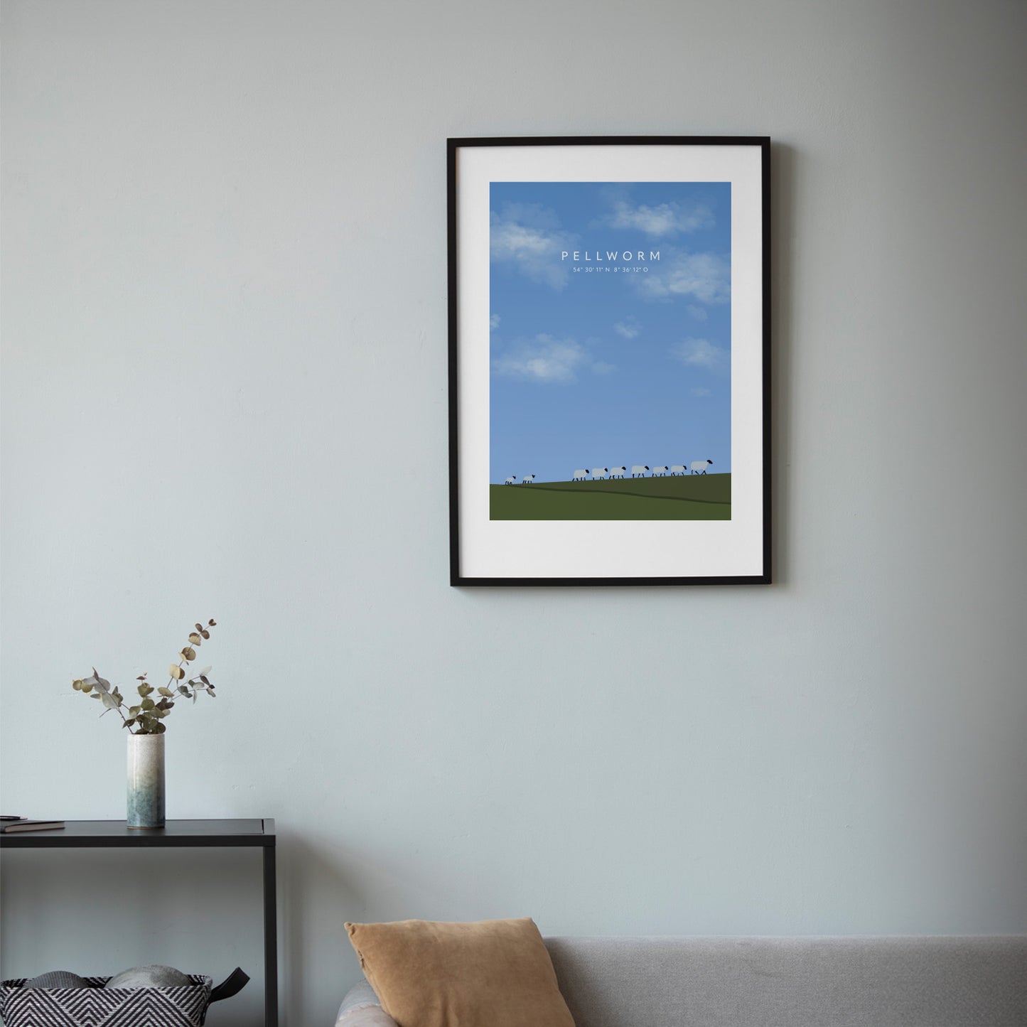 Minimalistisches Pellworm-Poster mit blauem Himmel, weißen Wolken, grünem Deich und stilisierten Schafen, gerahmt in Schwarz und an einer hellblauen Wand über einem Sofa aufgehängt, daneben eine schmale Konsole mit Vase und Zweigen.