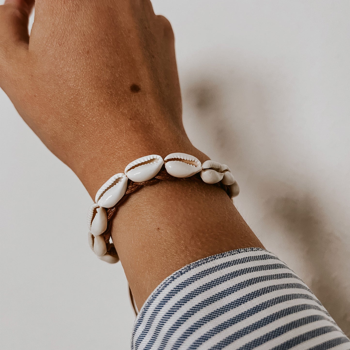 Armband aus weißen Kauri-Muscheln auf hellbraunem Band, getragen am Handgelenk vor natürlichem Hintergrund. Ausgesuchter Schmuck von Saltwaterdesigns mit Nordsee-Flair.