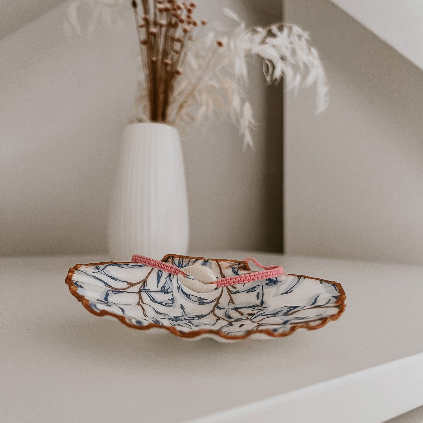 Kauri Muschelarmband in Rosa, präsentiert auf der Schmuckschale Muriwai von saltwaterdesigns. Auf weißem Schrank arrangiert, mit Blumen im Hintergrund – als Deko-Beispiel inszeniert.