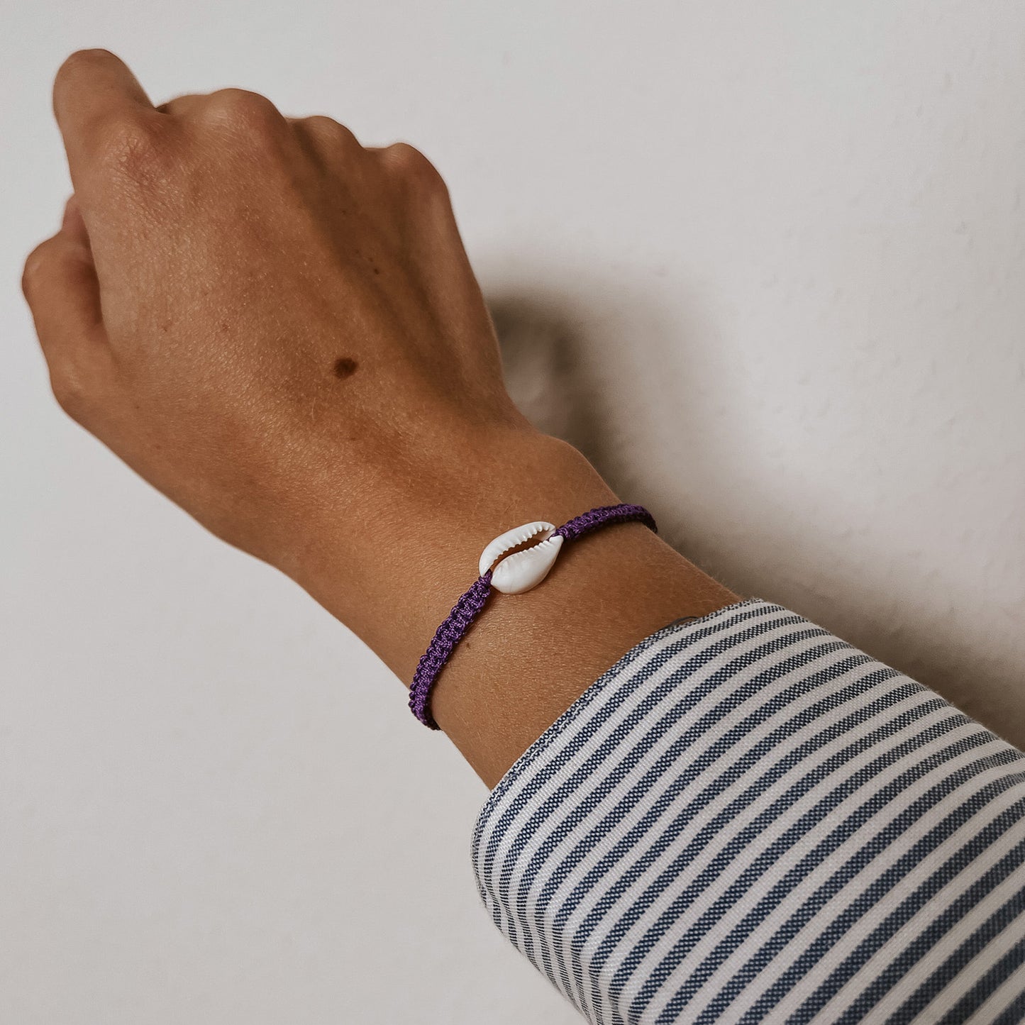 Geflochtenes Armband aus mit einer weißen Kauri Muschel in der Farbe lila. Getragen an einem Handgelenk. Bali Armband von saltwaterdesigns.