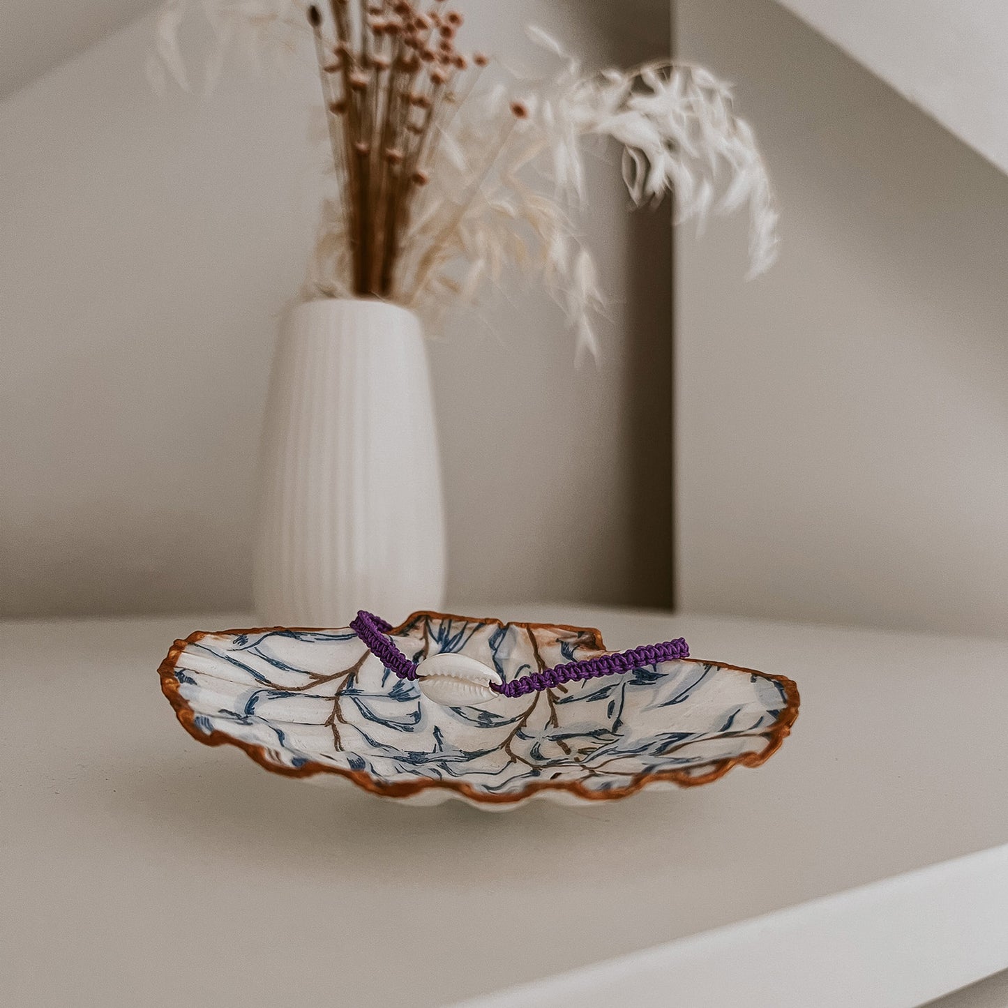 Lila Bali Muschelarmband, dekorativ platziert auf der Schmuckschale Muriwai von saltwaterdesigns. Die Szene auf einem weißen Schrank, umgeben von Blumen, zeigt eine natürliche und stilvolle Deko-Inspiration.