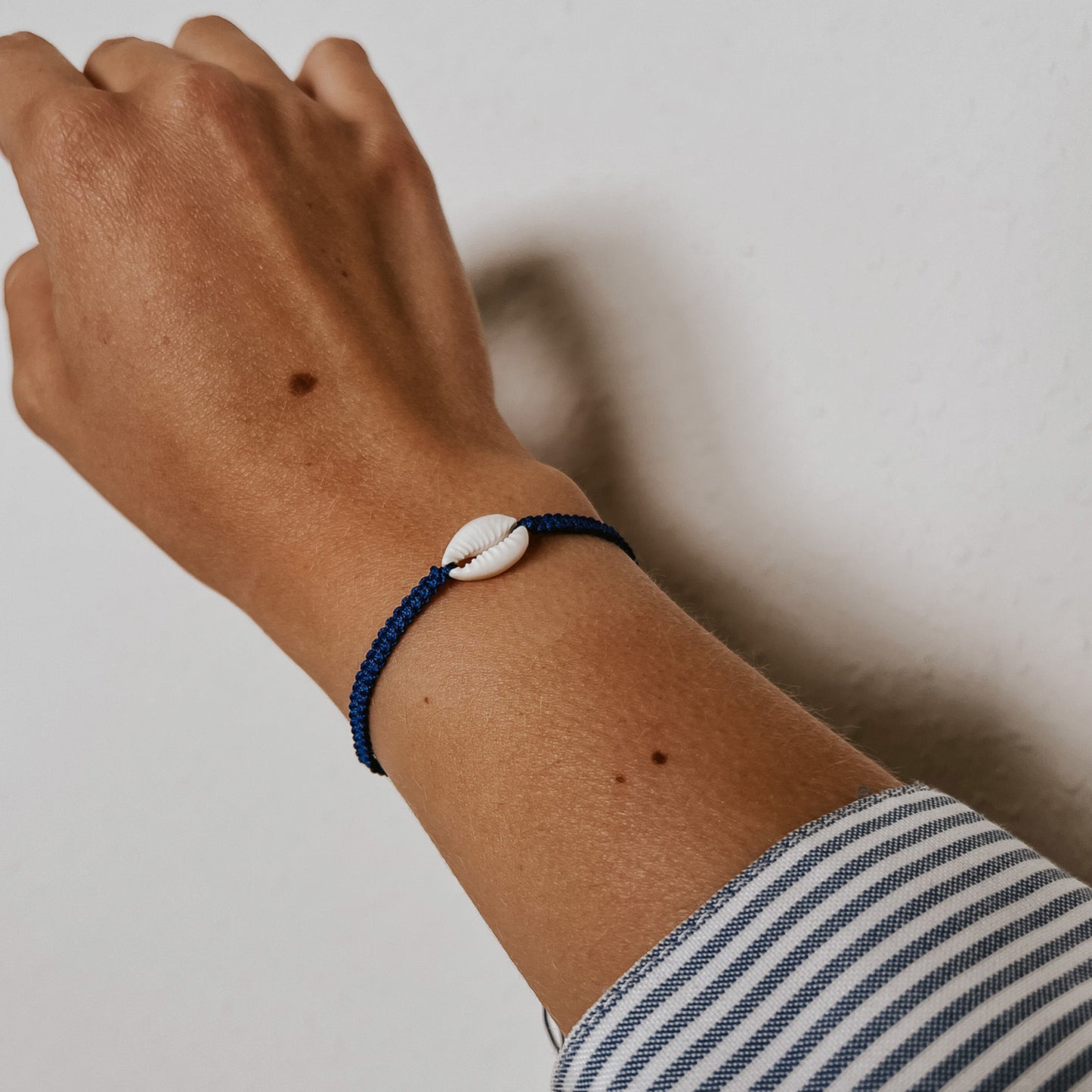 Bali Muschelarmband mit einer weißen Kauri-Muschel in der Mitte auf blauem Band, getragen am Handgelenk vor weißem Hintergrund. Handgemachter Schmuck von Saltwaterdesigns mit Küstenflair.