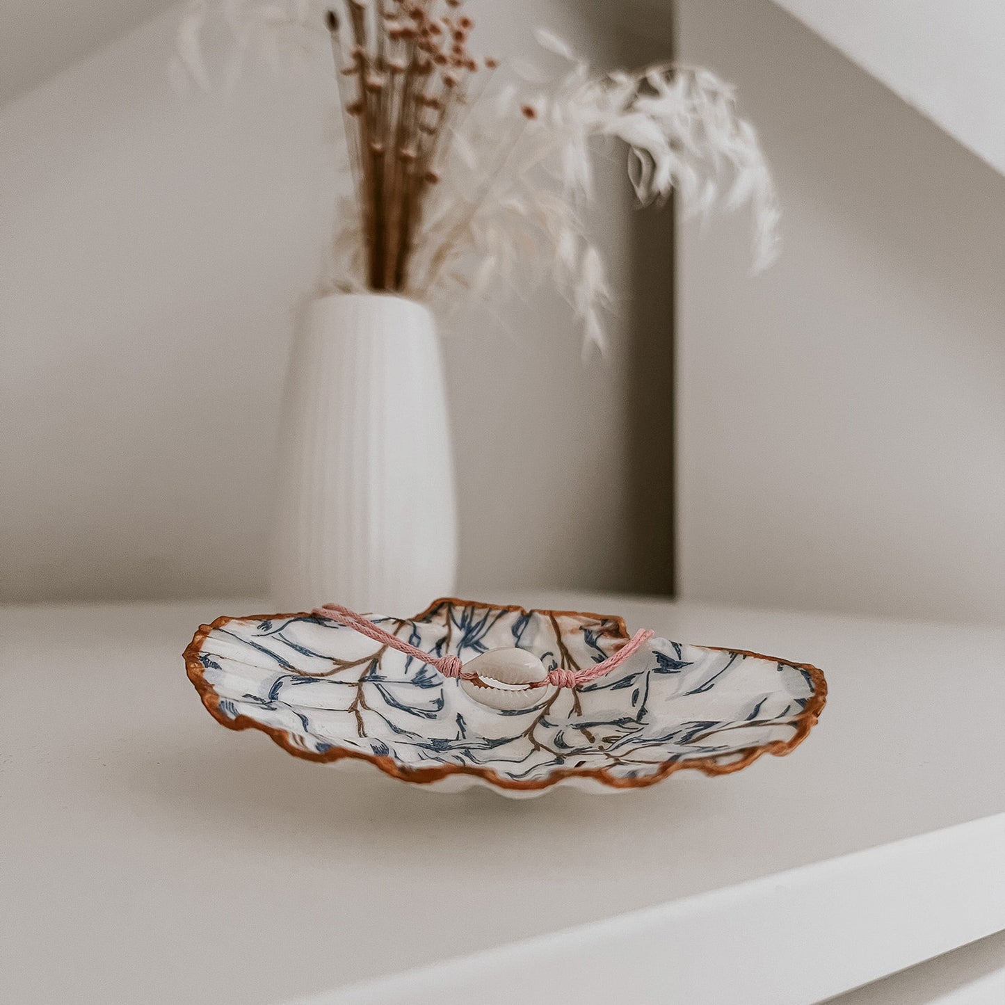 Zartes Ocean Kauri Muschelarmband in Rosa, dekorativ auf der Schmuckschale Muriwai von saltwaterdesigns präsentiert. Auf einem weißen Schrank arrangiert, mit Blumen im Hintergrund – ein elegantes Beispiel für stilvolle Wohn- und Dekoideen.