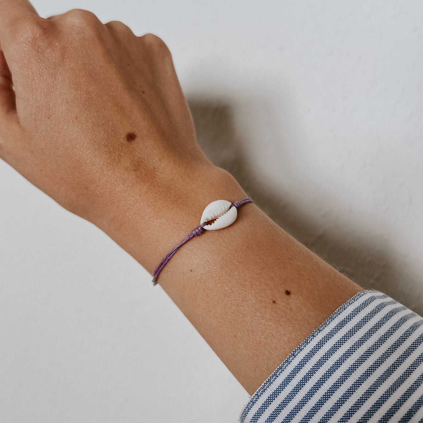 Zartes Ocean Armband mit einer weißen Kauri-Muschel auf lila Band, dezent am Handgelenk getragen. Das minimalistische Design von Saltwaterdesigns verleiht einen eleganten, maritimen Akzent für jeden Tag.