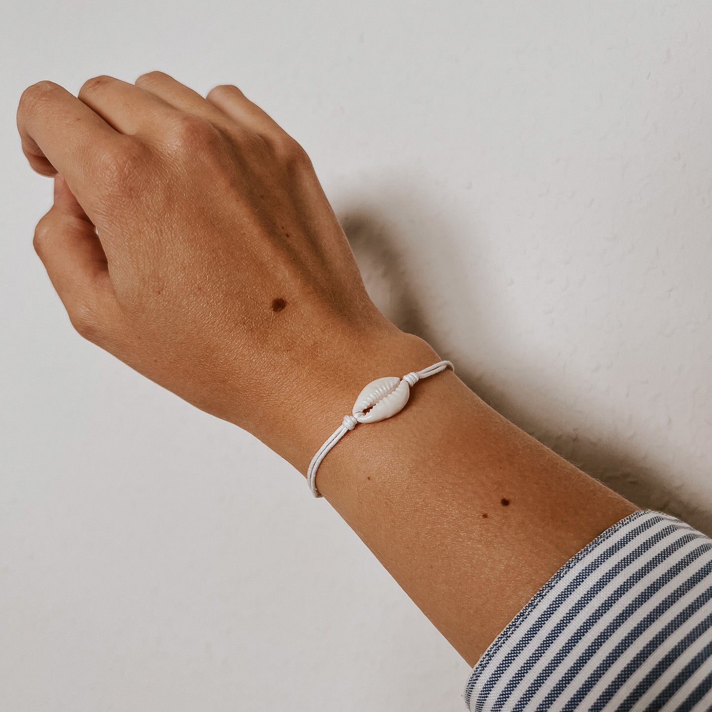 Feines Ocean Armband mit einer weißen Kauri-Muschel auf weißem Band, lässig am Handgelenk getragen. Das minimalistische Design von Saltwaterdesigns bringt maritimes Flair in den Alltag und wirkt besonders elegant und dezent.