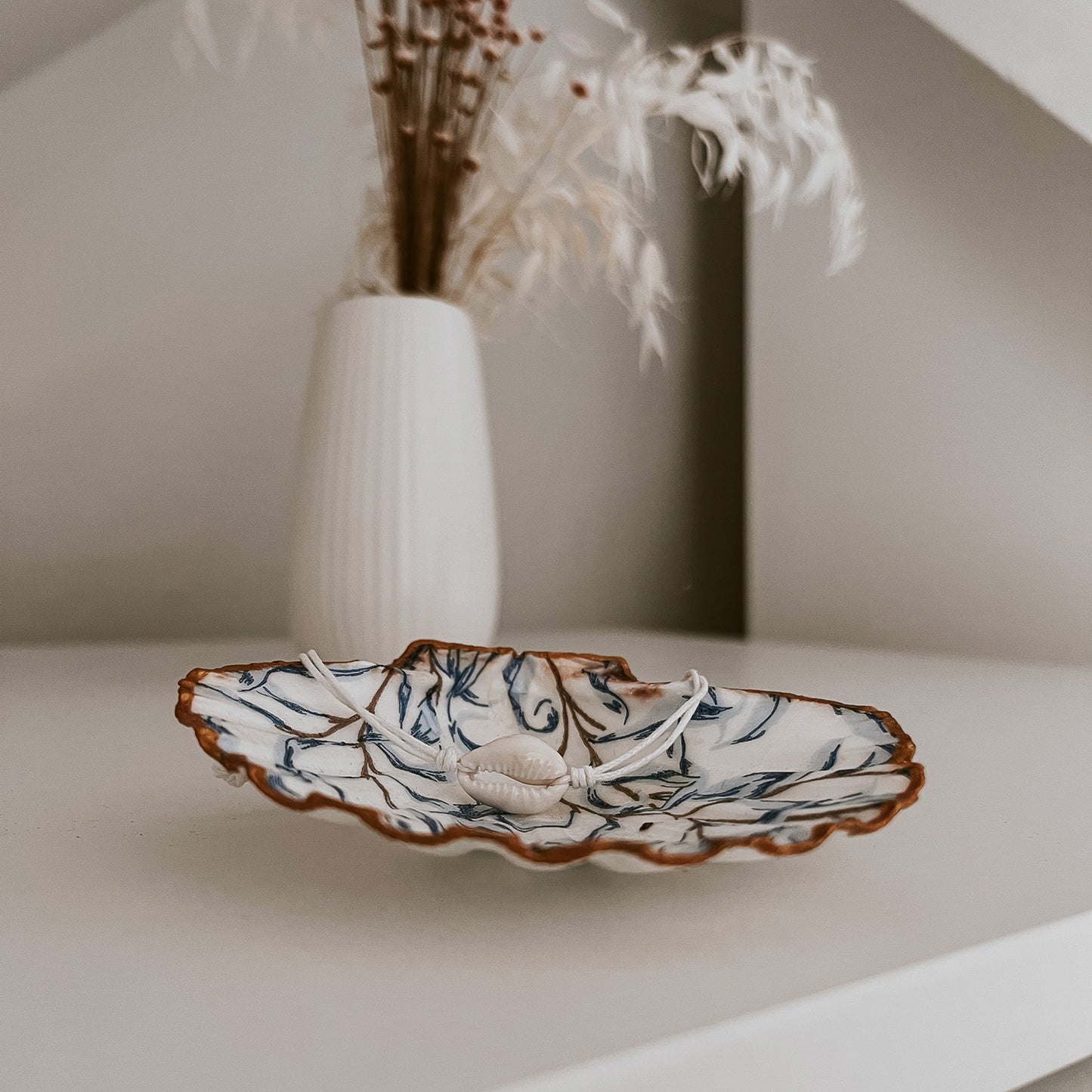 Feines Ocean“Kauri Muschelarmband in weiß, mittig auf der Schmuckschale Muriwai von saltwaterdesigns drapiert. Die Szene auf einem hellen Schrank, umgeben von zarten Blumen, zeigt ein stilvolles und maritimes Deko-Arrangement.