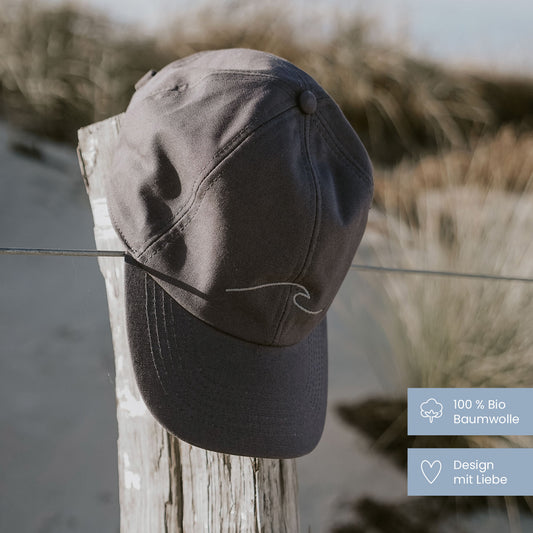 Dunkelgraue Cap mit gestickter Welle hängt auf einem verwitterten Holzpfahl am Dünenstrand, mit Gräsern und Meer im Hintergrund.