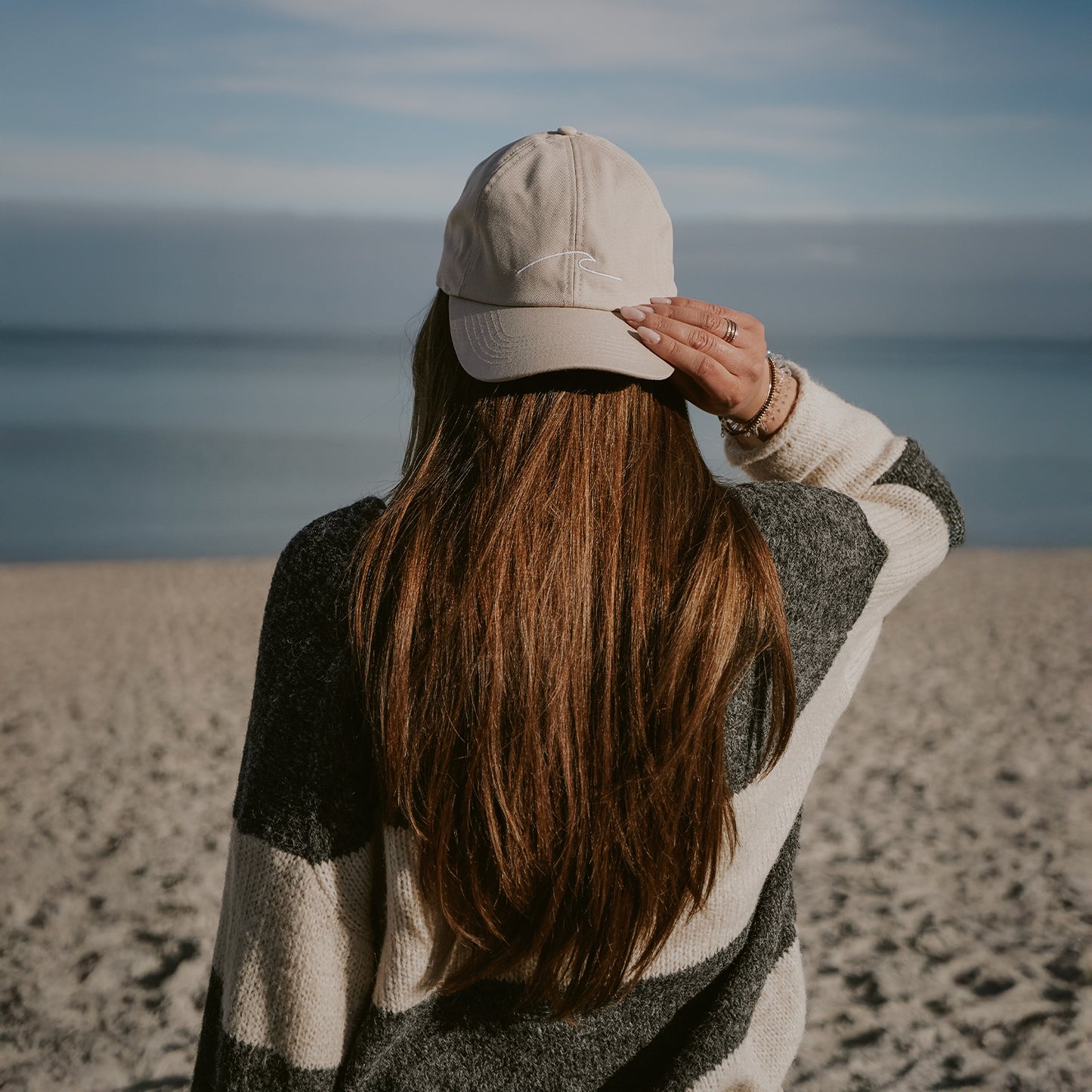 Person mit langen braunen Haaren, die eine beigefarbene Baseballkappe mit Wellenstickerei trägt, steht am Strand und blickt aufs Meer, während sie mit einer Hand den Schirm der Kappe hält.