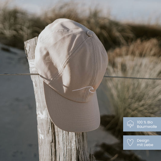 Beige Baseballkappe mit weißem Wellenlogo hängt an einem alten Holzpfosten, der mit einem Draht umzäunt ist. Im Hintergrund unscharfe Dünenlandschaft mit Gras. Unten rechts zwei grafische Labels: '100 % Bio Baumwolle' mit Baumwoll-Icon und 'Design mit Liebe' mit Herz-Icon.