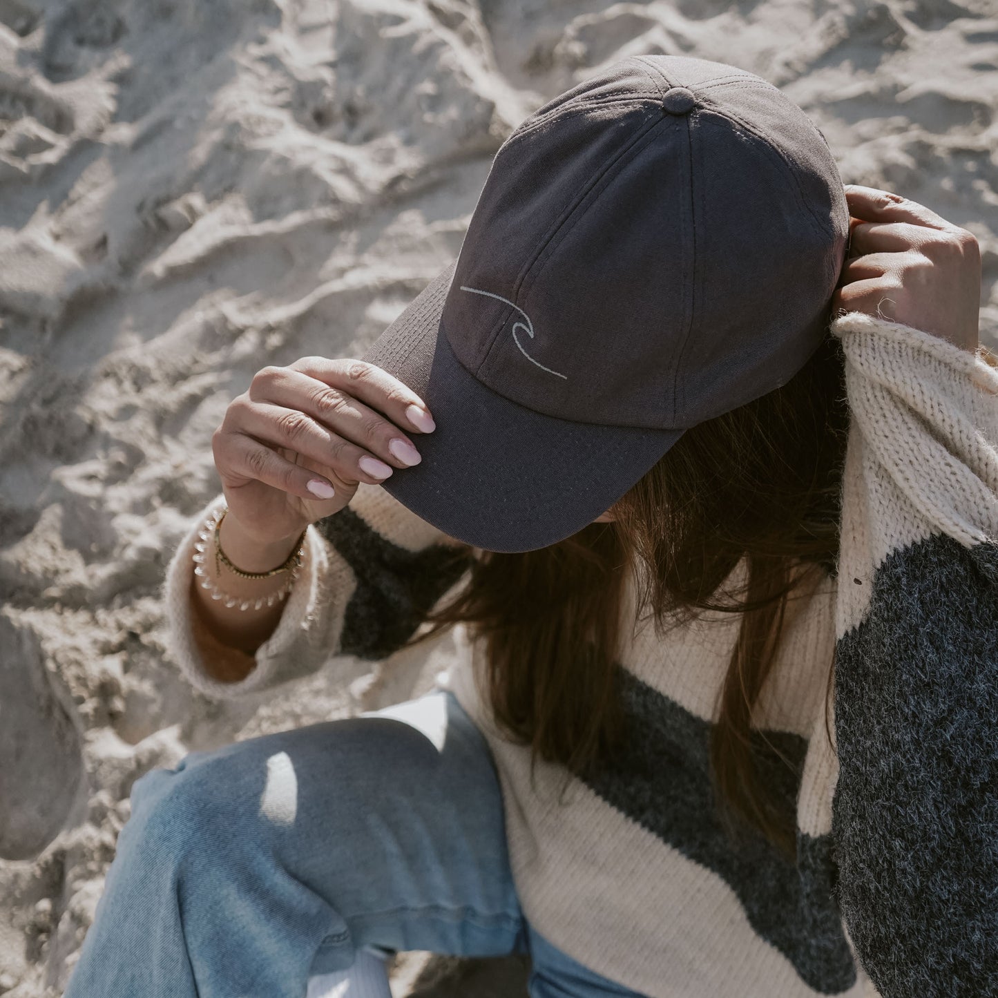 Frau mit dunkelgrauer Cap mit gestickter Welle sitzt am Sandstrand, trägt Strickpullover mit Blockstreifen und helle Jeans. Fokus auf die Cap und ihre Hand, die den Schirm hält.